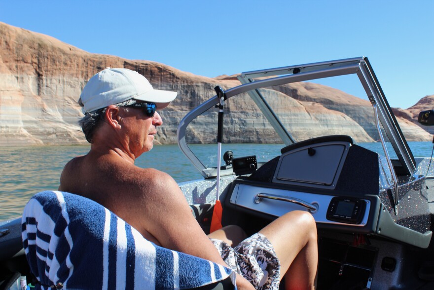 A shirtless man with sunglasses and a baseball cap sits on a seat in an open-air boat on a lake surrounded by orange and white rock walls