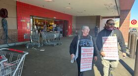 Two people with white signs hanging from their necks explain that they are striking. They're standing near a parking lot in a shopping center.